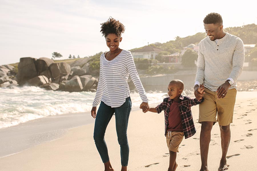 Personal Insurance - Family Smiles as They Walk Along the Beach on a Misty Afternoon, Parents Holding Their Young Son's Hands, Rocks and Surf Behind Them