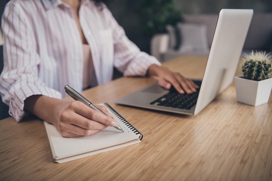 Resources - Woman Makes Notes in a Book While Using Her Laptop in a Home Office, a Small Cactus on Her Desk